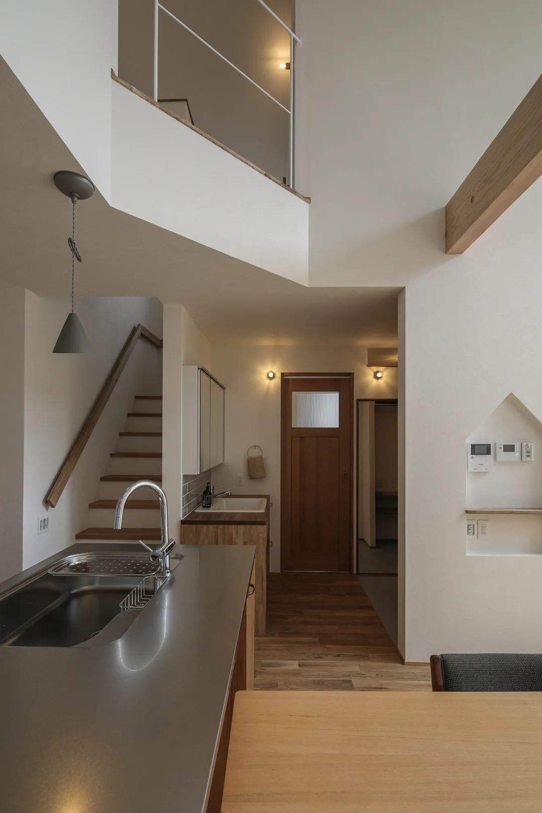 左手前にキッチン、右手前にダイニングテーブル、正面奥に木製のドア。上部は吹き抜けていて、2階廊下の白い手すりが見える。