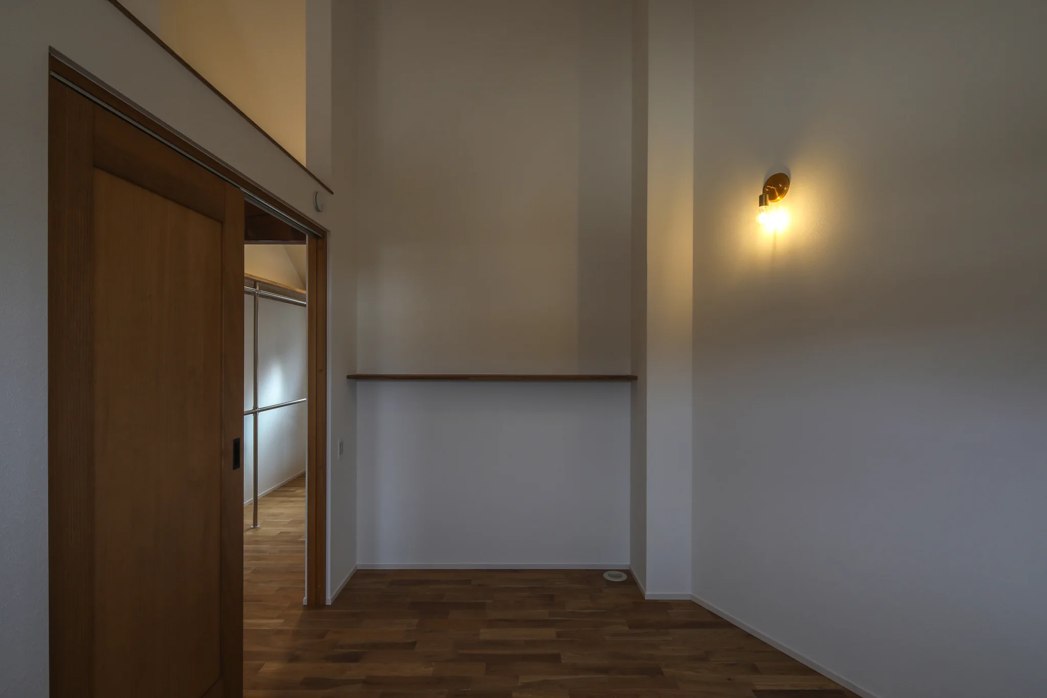 主寝室。床が台形をしている。斜め部分の壁には壁付けの照明がある。左手には入口の引き戸。茶色い木製。正面には飾り棚がある。床は無垢のオークのフローリング