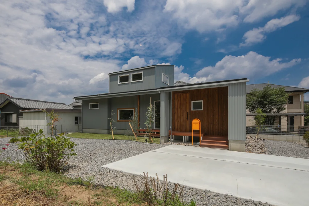 青空の屋外、中心にグレーの壁の一戸建て。壁の一部に木が貼られている。2階部分が小さい。黄色いポストが玄関ドアのそばにある。手前には駐車スペースの土間と砂利、芝生がある