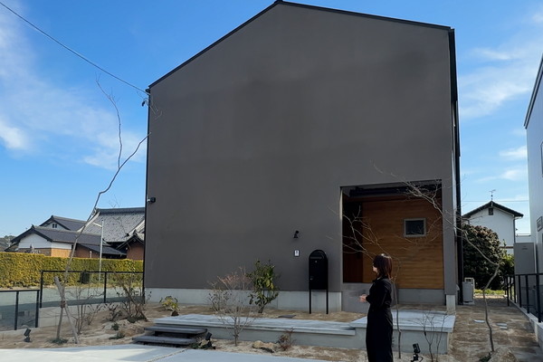 ブラウンの壁をした三角屋根の家の前に、解説するために設計士が立っている